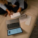 Kobieta na łóżku przy laptopie porządkuje przelewy i dokumenty finansowe